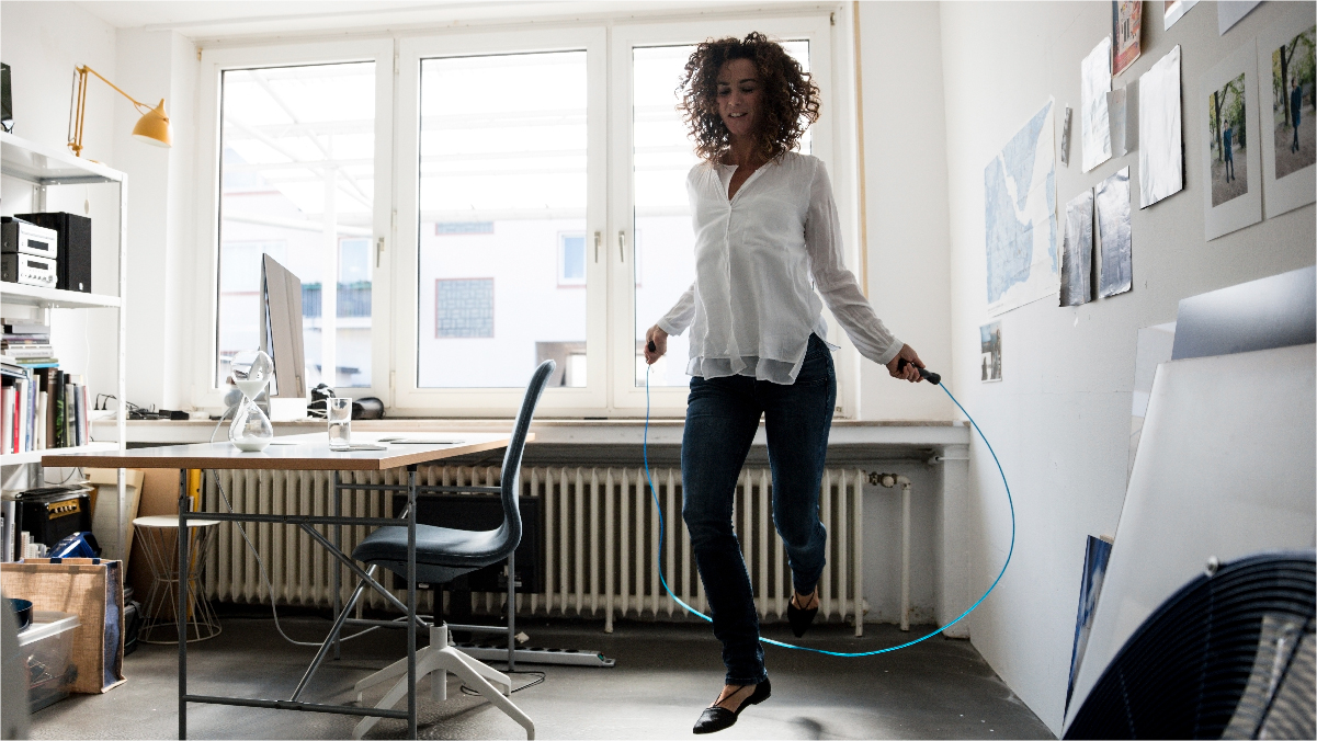 A woman is in her home office, dressed in work attire and is taking a Stress Care beak by doing some jump rope.
